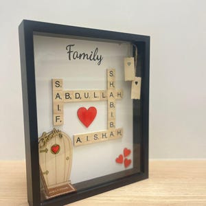 May include: A framed family name display in a black frame. The word "Family" is written at the top. Wooden letter tiles spell out names, with a red heart in the center. A wooden door with a heart and a "Welcome" mat are also included.