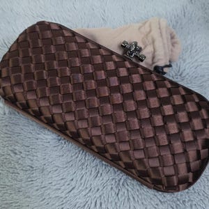 May include: A rectangular dark brown clutch bag featuring a woven satin ribbon exterior. The bag has a metallic gunmetal-toned clasp closure at the top and is accompanied by a soft beige fabric drawstring storage pouch.