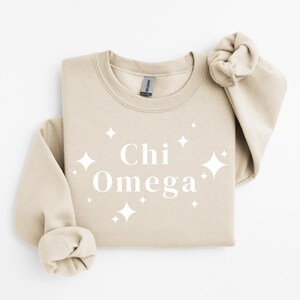 Könnte beinhalten: Beiges Sweatshirt mit Rundhalsausschnitt und dem weißen Schriftzug "Chi Omega", umgeben von weißen Sternen. Das Sweatshirt ist gefaltet, ein Ärmel ist sichtbar. Das Kleidungsstück ist aus weichem Material.
