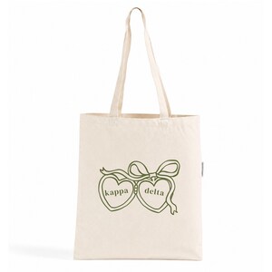 Puede incluir: Una bolsa de tela de color natural con dos contornos de corazón en verde oliva unidos por un lazo. El corazón izquierdo contiene la palabra "kappa" y el corazón derecho la palabra "delta" en una fuente serif sencilla en minúsculas.