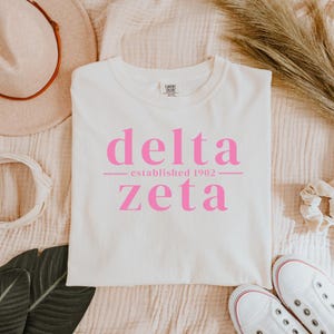 Puede incluir: Camiseta de color crema con las palabras "delta" y "zeta" en rosa, con "established 1902" debajo. La camiseta está sobre una superficie texturizada, con un sombrero, zapatos y elementos decorativos en el fondo.