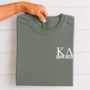 Peut inclure: Un t-shirt à col rond vert sauge avec les lettres grecques blanches KA et le texte KAPPA DELTA imprimé sur le côté inférieur droit. Le t-shirt possède une étiquette de marque Comfort Colors à l'intérieur du col avec la taille M imprimée.