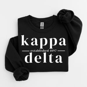 Puede incluir: Una sudadera negra de manga larga con cuello redondo que presenta texto blanco que dice kappa delta con las palabras established 1897 centradas entre las dos líneas de texto. La prenda tiene cuello, puños y dobladillo acanalados con etiqueta Gildan.