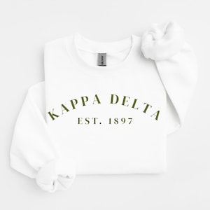 Könnte beinhalten: Ein weißes Sweatshirt mit Rundhalsausschnitt und dem Text KAPPA DELTA in olivgrüner Schrift, der über dem Text EST. 1897 gebogen ist. Das Kleidungsstück hat lange Ärmel mit gerippten Bündchen und ein Gildan-Markenetikett am Innenkragen.
