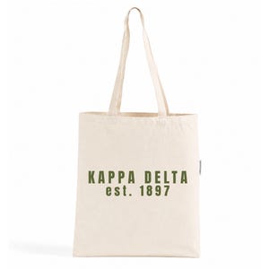 Op de afbeelding: Een crèmekleurige canvas draagtas met twee lange schouderbanden. De voorkant van de tas is voorzien van de donkergroene tekst KAPPA DELTA est. 1897, gedrukt in een vet, schreefloos lettertype. De tas heeft een rechthoekige vorm en plat design.