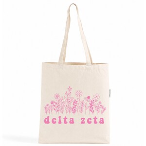 Peut inclure: Un sac fourre-tout en toile couleur crème doté de deux longues anses et d'un imprimé graphique rose. Le motif présente une collection de fleurs dessinées au trait au-dessus du texte en minuscules "delta zeta" en police rose arrondie.