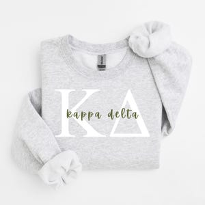 Könnte beinhalten: Ein hellgraues Sweatshirt mit Rundhalsausschnitt, das große weiße griechische Buchstaben K und Delta zeigt, mit den Worten kappa delta in einer kleineren olivgrünen Schreibschrift in der Mitte. Das Kleidungsstück hat lange Ärmel und gerippte Bündchen.