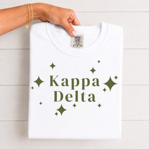 Peut inclure: Un t-shirt blanc à manches courtes avec le texte vert foncé Kappa Delta centré sur la poitrine. Le motif est entouré de diverses étoiles à quatre branches vert foncé. Le t-shirt porte une étiquette de marque Comfort Colors à l'encolure.