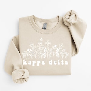 Puede incluir: Una sudadera beige de cuello redondo con un gráfico de líneas blancas de varias flores silvestres en el pecho. Debajo del diseño floral, el texto "kappa delta" está impreso en una fuente blanca redondeada de estilo retro. Tiene mangas largas.