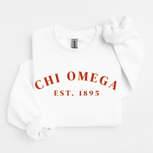 Puede incluir: Una sudadera blanca con el texto "CHI OMEGA EST. 1895" en letras rojas con contorno dorado. La sudadera tiene cuello redondo y mangas largas.