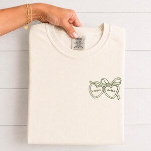 Pode incluir: Uma t-shirt de manga curta na cor creme com um gráfico verde escuro no lado inferior direito. O gráfico mostra dois medalhões em forma de coração conectados por um laço, com o texto kappa dentro do coração esquerdo e delta no direito.