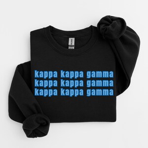 Op de afbeelding: Zwart sweatshirt met de tekst "kappa kappa gamma" in blauw, drie keer gestapeld. Het sweatshirt is opgevouwen, met de mouwen en kraag zichtbaar. De stof lijkt zacht.