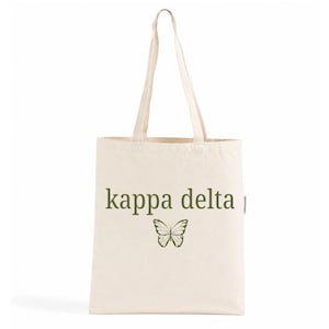 Puede incluir: Una bolsa de tela color crema con el texto kappa delta impreso en verde oliva. Debajo del texto hay un gráfico detallado de una mariposa en verde oliva. La bolsa tiene dos asas largas para el hombro y un cuerpo rectangular de tela tejida.