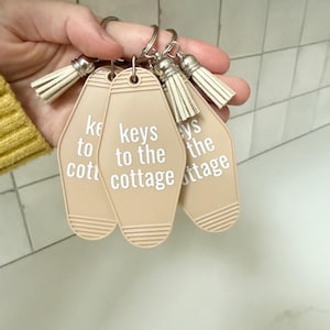 May include: A set of three beige keychains with the text "keys to the cottage" in white. Each keychain has a silver ring and a cream-colored tassel. The keychains are held in a hand.