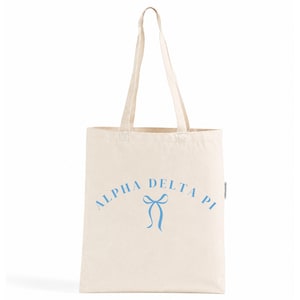 Puede incluir: Un bolso de tela de color natural con dos asas largas para el hombro. La parte frontal presenta el texto ALPHA DELTA PI impreso en una fuente sans-serif azul claro, dispuesto en un arco sobre un gráfico azul claro de un lazo de cinta.