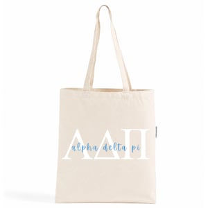 Op de afbeelding: Een natuurkleurige canvas draagtas met de witte Griekse letters Alpha Delta Pi en de tekst alpha delta pi in lichtblauw cursief schrift in het midden gedrukt. De tas heeft twee lange schouderbanden en een verticaal rechthoekig ontwerp.
