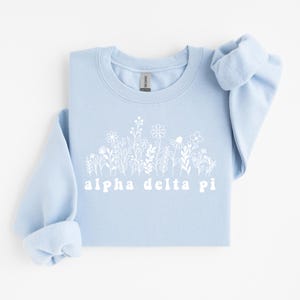 Op de afbeelding: Een lichtblauw sweatshirt met een wit bloemenontwerp en de tekst "alpha delta pi". De sweatshirt heeft een ronde hals en lange mouwen met opgerolde manchetten. Het ontwerp toont verschillende bloemen en bladeren.