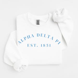 May include: A white crewneck sweatshirt with the light blue text "ALPHA DELTA PI EST. 1851" across the chest. The sweatshirt has long sleeves and a classic, casual design, perfect for everyday wear.