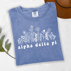 Puede incluir: Una camiseta azul con un diseño floral blanco y el texto "alpha delta pi". La camiseta está hecha de un material suave y tiene cuello redondo. La etiqueta dice "Comfort Colors".