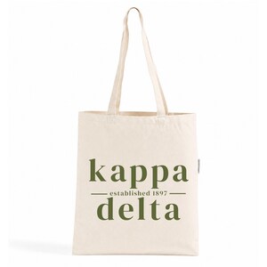 Puede incluir: Un bolso de tela color crema con dos asas largas para el hombro. La parte frontal presenta un texto en verde oliva oscuro que dice "kappa" en la línea superior, "established 1897" entre dos líneas horizontales y "delta" abajo.