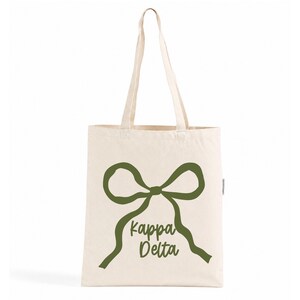 Pode incluir: Uma sacola de lona na cor creme com duas alças longas e uma estampa gráfica em verde oliva escuro na frente. O design apresenta um laço grande com o texto Kappa Delta escrito em uma fonte cursiva logo abaixo do laço.