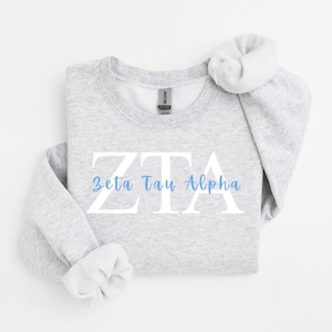 Op de afbeelding: Een lichtgrijze sweater met ronde hals voorzien van de grote witte letters ZTA met de woorden Zeta Tau Alpha gedrukt in een lichtblauw scriptlettertype in het midden. Het kledingstuk heeft lange mouwen met geribbelde boorden en een standaard hals.