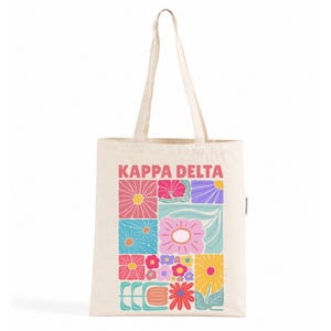 Könnte beinhalten: Eine cremefarbene Stofftasche aus Segeltuch mit dem Schriftzug KAPPA DELTA in rosa Buchstaben über einem Raster aus bunten Blumenillustrationen. Das Design zeigt verschiedene stilisierte Blumen in Rosa, Blau, Gelb, Lila und Türkis.
