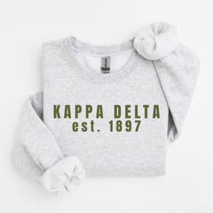 Könnte beinhalten: Ein hellgraues Sweatshirt mit Rundhalsausschnitt und dem dunkel olivgrünen Schriftzug KAPPA DELTA est. 1897 auf der Brust. Das Kleidungsstück hat lange Ärmel mit gerippten Bündchen und einen gerippten Halsausschnitt sowie ein Gildan-Etikett am Kragen.
