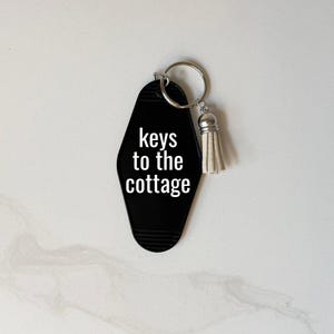 May include: Black motel-style keychain with the words "keys to the cottage" in white. The keychain has a silver key ring and a small, cream-colored tassel. The background is a light, neutral color.