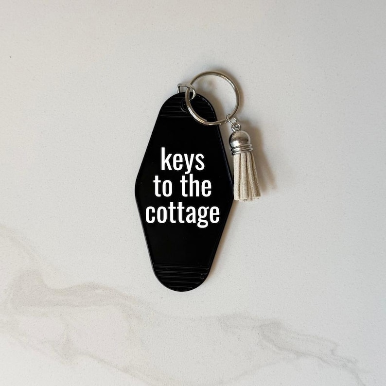 May include: Black motel-style keychain with the words "keys to the cottage" in white. The keychain has a silver key ring and a small, cream-colored tassel. The background is a light, neutral color.