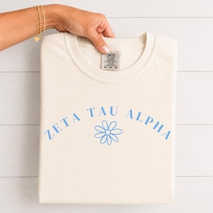 Op de afbeelding: Een crèmekleurig t-shirt met korte mouwen met de tekst ZETA TAU ALPHA in een lichtblauw gebogen lettertype boven een eenvoudige lichtblauwe madeliefjesafbeelding. Het t-shirt heeft een Comfort Colors-label bij de hals met maat M.