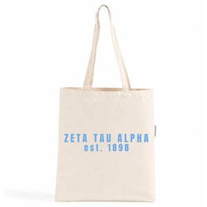 Op de afbeelding: Een crèmekleurige canvas draagtas met twee lange schouderbanden. De voorzijde van de tas is voorzien van de tekst "ZETA TAU ALPHA" in een lichtblauw schreefloos lettertype, met de tekst "est. 1898" er direct onder in dezelfde blauwe kleur.
