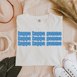 Könnte beinhalten: Ein weißes T-Shirt mit dem Text "kappa kappa gamma" in Blau, dreimal wiederholt. Das Shirt besteht aus einem weichen Material. Das Shirt wird auf einer hellrosa Oberfläche präsentiert. Wahrscheinlich ein Comfort Colors-Shirt.