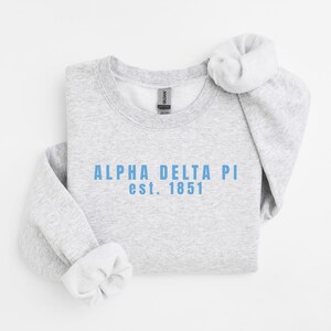 Puede incluir: Una sudadera gris claro con el texto "ALPHA DELTA PI est. 1851" en azul claro. La sudadera tiene cuello redondo y mangas largas con puños enrollados. La etiqueta de la marca Gildan es visible.
