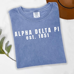 Puede incluir: Una camiseta azul Comfort Colors con el texto blanco "ALPHA DELTA PI est. 1851". La camiseta está doblada y sobre una superficie blanca. Un sombrero marrón es visible en el fondo.
