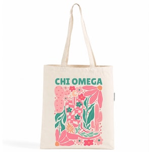 Puede incluir: Un bolso de tela color crema con el texto CHI OMEGA en color verde azulado. El diseño gráfico incluye una bota vaquera a cuadros rosas y blancos, varias flores rosas y rojas, hojas verdes, un cactus de pera espinosa rosa y un cactus alto.
