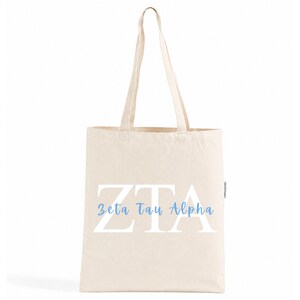 Op de afbeelding: Een natuurkleurige canvas draagtas met twee lange schouderbanden. De voorzijde is voorzien van grote witte blokletters met de tekst ZTA, waarbij de woorden Zeta Tau Alpha in een lichtblauw cursief lettertype over de letters zijn gedrukt.