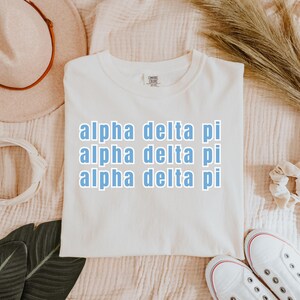 Peut inclure: T-shirt crème avec l'inscription "alpha delta pi" en bleu clair, répétée trois fois. Le t-shirt est en matière douce et est présenté sur une surface texturée. Des baskets blanches avec des accents rouges sont visibles en bas à droite.
