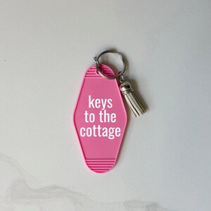 May include: A pink motel-style keychain with the words "keys to the cottage" in white. The keychain has a silver key ring and a white tassel. The background is a light, neutral color.