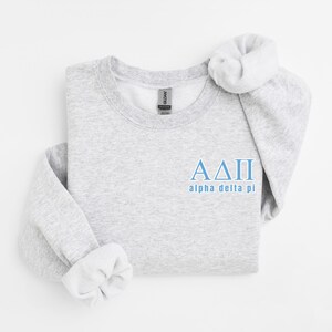 Könnte beinhalten: Ein hellgraues Sweatshirt mit den griechischen Buchstaben ΑΔΠ in Hellblau und den Worten "alpha delta pi" darunter. Das Sweatshirt hat einen Rundhalsausschnitt und lange Ärmel, mit einem weißen Scrunchie auf der Schulter.