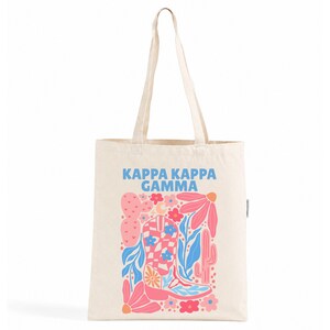 Peut inclure: Un sac fourre-tout en toile couleur crème avec le texte bleu KAPPA KAPPA GAMMA au-dessus d'une impression graphique colorée. Le motif comprend une botte de cow-boy à carreaux, des cactus roses, des feuilles bleues et des fleurs roses et rouges.