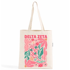 Op de afbeelding: Een crèmekleurige canvas tas met de tekst DELTA ZETA in roze letters. Het grafische ontwerp bevat een roze en wit geblokte cowboylaars, roze bloemen, groene bladeren, een roze schijfcactus en een hoge roze cactus.