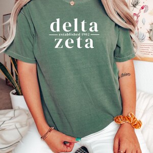 Puede incluir: Una camiseta verde con las palabras "delta" y "zeta" en blanco, con "established 1902" en medio. La camiseta es de color liso y tiene mangas cortas. La persona lleva una goma de pelo dorada y pulseras.