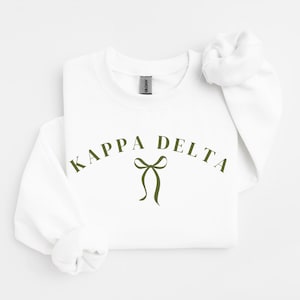 Puede incluir: Una sudadera blanca de cuello redondo con el texto KAPPA DELTA en una fuente arqueada de color verde oliva, centrada sobre un gráfico de lazo simple en verde oliva. La prenda tiene mangas largas con puños acanalados y una etiqueta Gildan.