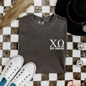 Könnte beinhalten: Ein anthrazitfarbenes T-Shirt mit den griechischen Buchstaben XΩ und den Worten "chi omega" in Weiß. Das Shirt liegt gefaltet auf einer karierten Oberfläche, mit weißen Turnschuhen und einem schwarzen Hut im Hintergrund.