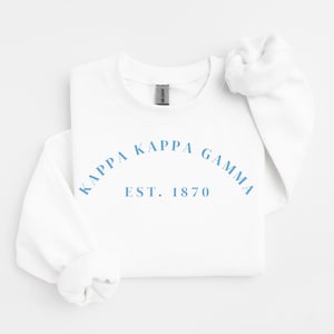Peut inclure: Un sweat-shirt blanc avec l'inscription "KAPPA KAPPA GAMMA EST. 1870" en bleu clair, disposée en arc de cercle. Le sweat-shirt a des manches longues et un col rond. L'étiquette de la marque Gildan est visible.