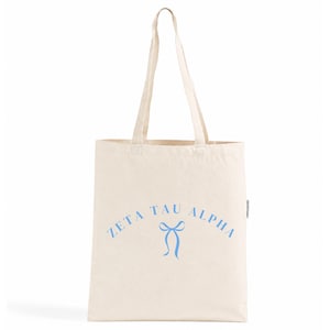 Pode incluir: Uma sacola de lona na cor creme com duas alças longas. A parte frontal da bolsa exibe o texto "ZETA TAU ALPHA" em uma fonte azul clara, arqueado sobre um gráfico simples em azul claro de um laço de fita amarrado centralizado.