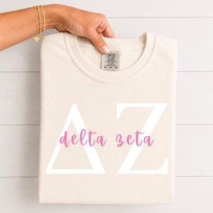 Puede incluir: Una camiseta de manga corta color crema con las letras griegas Delta y Zeta en blanco grande y las palabras delta zeta impresas en cursiva rosa en el centro. La camiseta tiene una etiqueta de la marca Comfort Colors en el cuello interior.