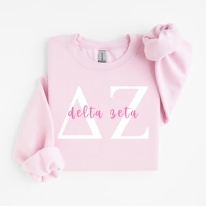 Peut inclure: Un sweat-shirt à col rond rose clair arborant les lettres grecques Delta et Zeta en blanc, avec les mots delta zeta imprimés en écriture cursive rose au centre. Le vêtement possède des manches longues ainsi qu'un col et des poignets côtelés.