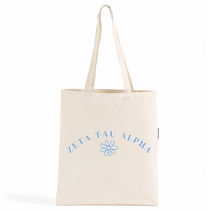 Könnte beinhalten: Eine cremefarbene Stofftasche aus Segeltuch mit zwei langen Schulterriemen. Auf der Vorderseite befindet sich der Schriftzug "ZETA TAU ALPHA" in einer hellblauen serifenlosen Schrift, bogenförmig über einer hellblauen Strichzeichnung einer Margerite.
